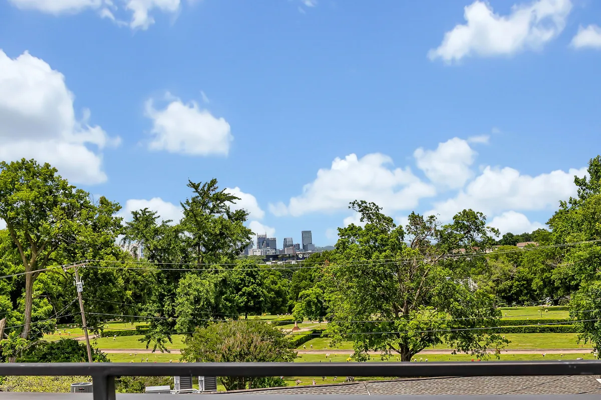 A lush green park scene with sprawling lawns, trees, and a distant city skyline under a bright blue sky. People can be seen walking, jogging, and relaxing on the grass, suggesting a lively and inviting public space ideal for outdoor activities.