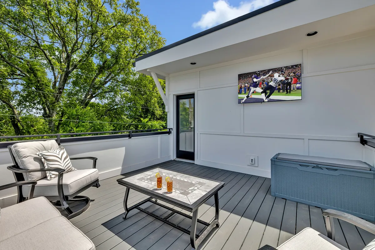 This image features a stylish rooftop outdoor seating area, complete with a flat-screen TV, a coffee table set with drinks, and lush greenery in the background. The arrangement of ample seating encourages social gatherings and relaxation, while the Smart TV serves as a focal point for entertainment.