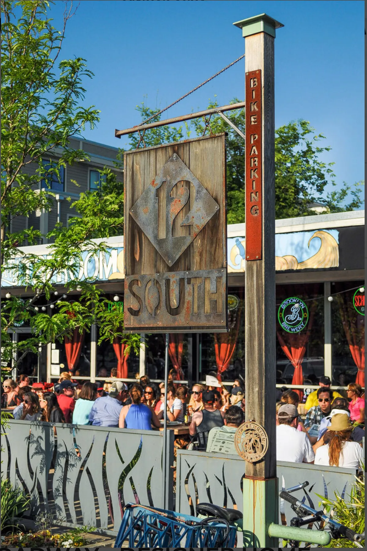 A bustling neighborhood streetscape with the distinctive 12 South sign, groups of people enjoying themselves, and visible bike parking. The scene buzzes with local energy and hints at walkable dining options.