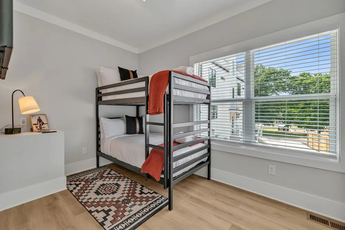 A playful yet cozy bunk bedroom with a sturdy bunk bed, an area rug, a lamp, and a window providing natural light and views. The room feels inviting for children or groups, with space for both rest and play.