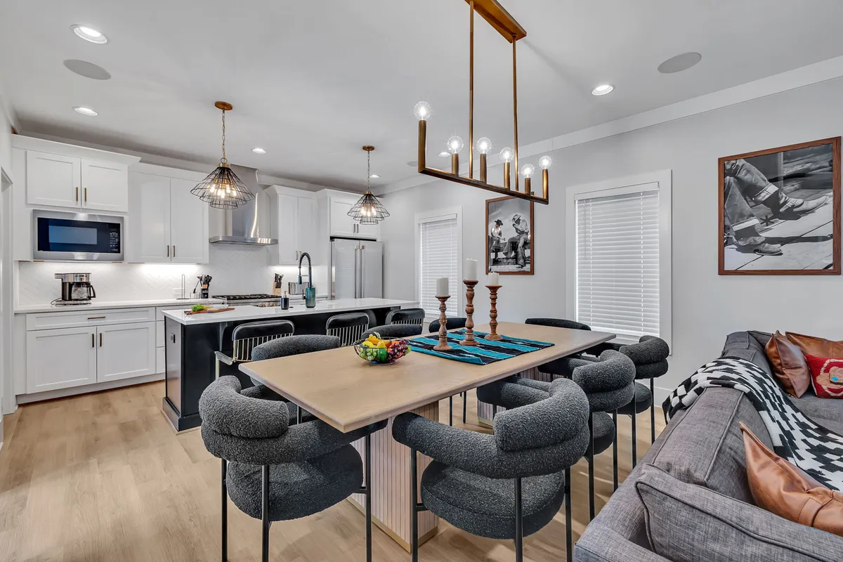 The image showcases an inviting open-concept living and dining area, with a warmly lit dining table adorned with candles, a stylish chandelier overhead, a modern kitchen island at the back, striking artwork on the wall, and a plush, comfortable sofa. The scene suggests a space perfect for gathering, sharing meals, and enjoying quality time.