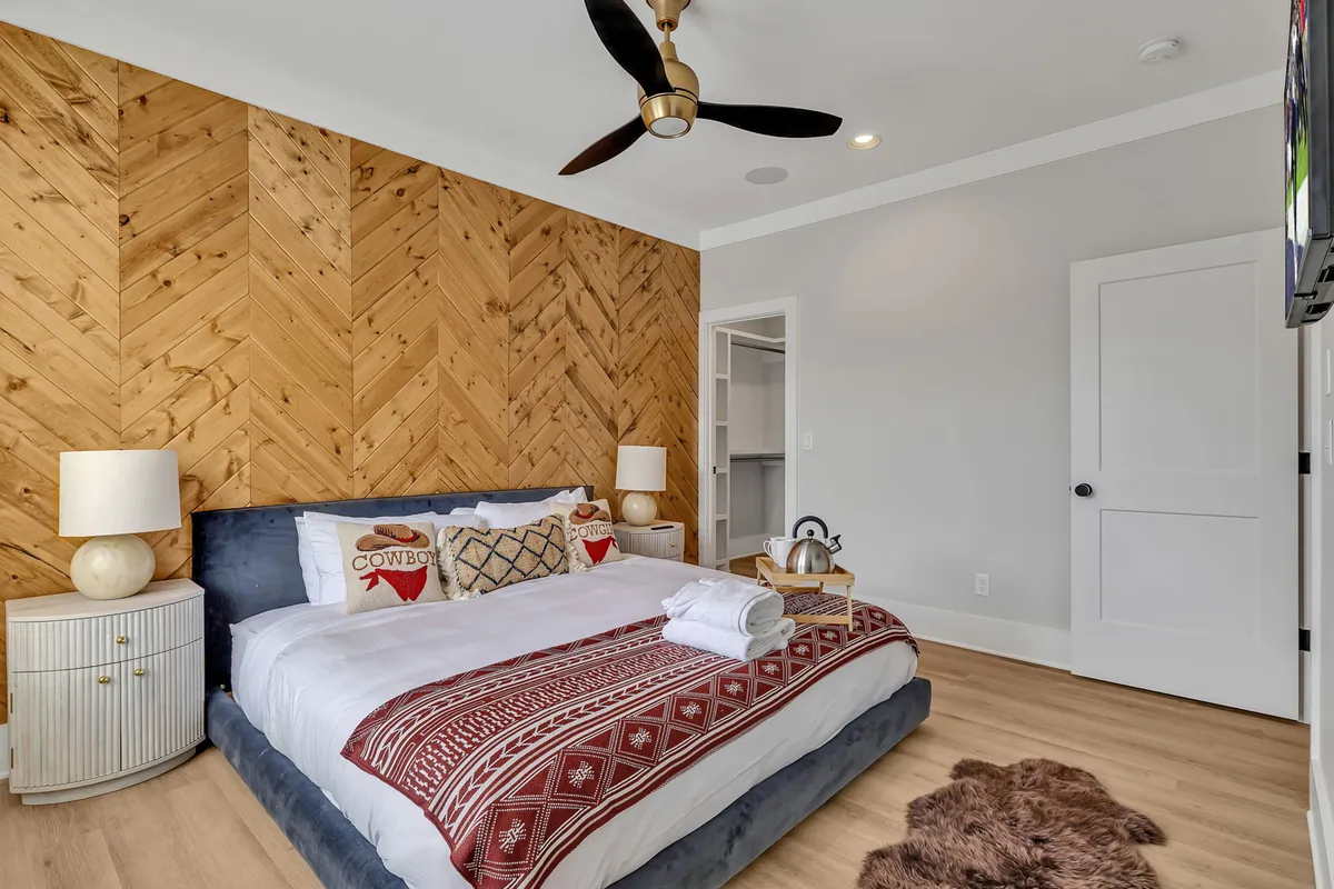 This image displays a spacious primary bedroom featuring a king-sized bed with decorative pillows, a wooden accent wall adding warmth, a nightstand with a lamp, and a soft rug on the floor. Soft natural light filters into the room, creating a serene and restful atmosphere.